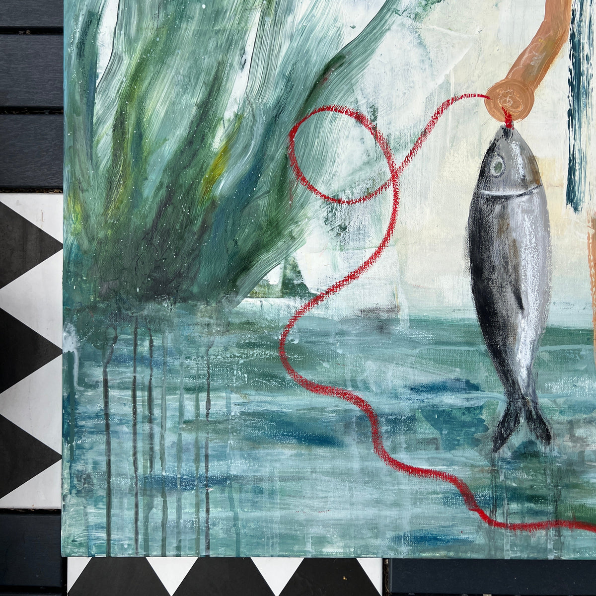 A close-up of a painted artwork showing a fish hanging from a red string, with green and blue water-like forms and a pale background. Part of a black-and-white checkered surface is visible around the artwork.