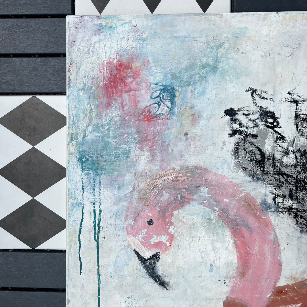 An abstract painting featuring a pink flamingo and black bird silhouettes on a textured white and blue background, displayed next to a black and white diamond-patterned wooden panel.