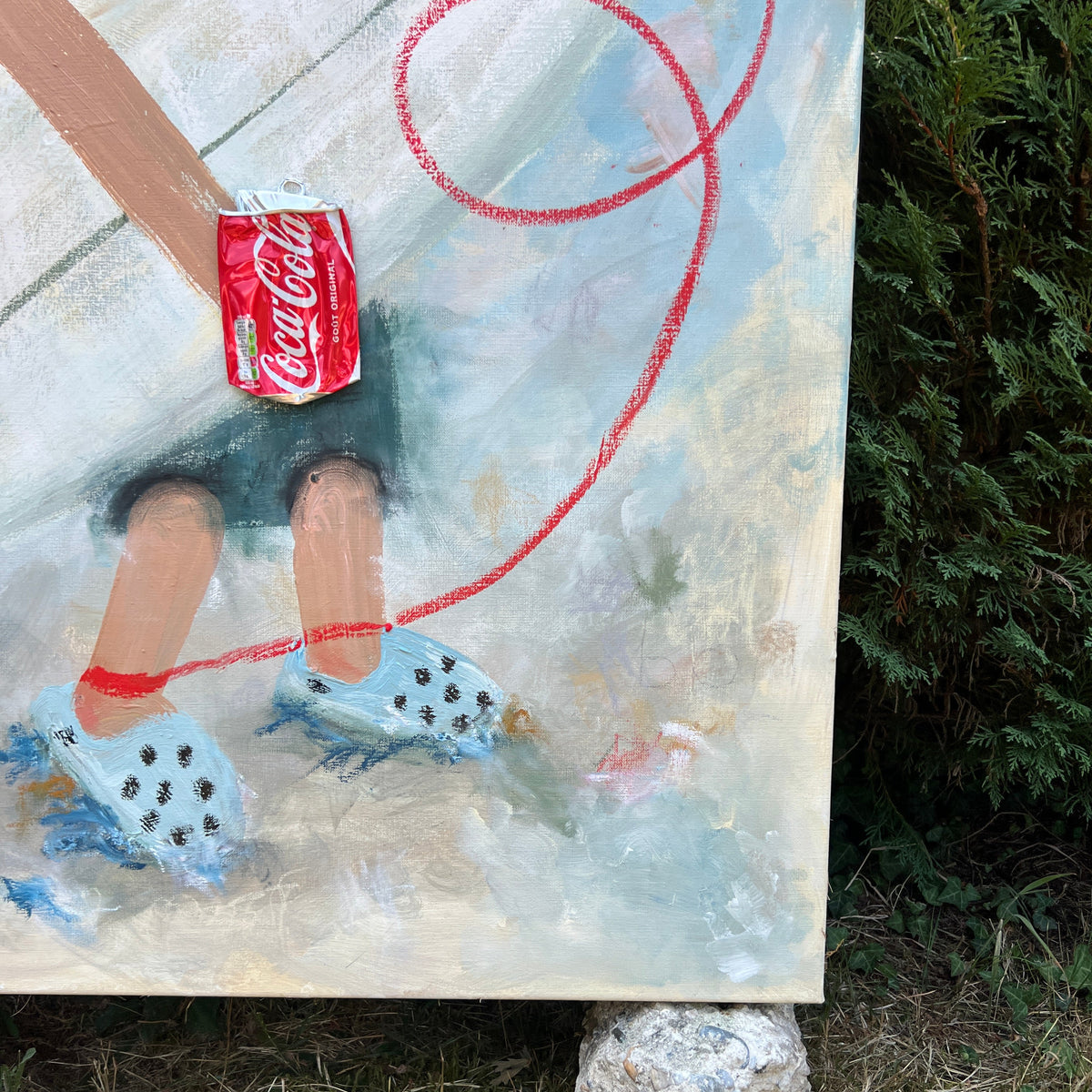 A close-up of a painted canvas showing a stylized figure holding a red Coca-Cola can, with blue-and-white dice and a red looped line, photographed outdoors on grass with a rock underneath the canvas.