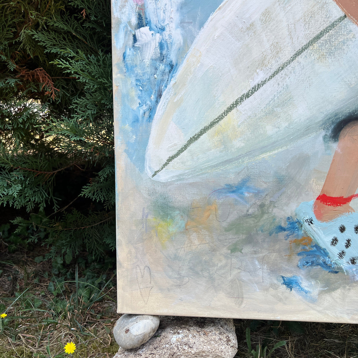A large canvas painting depicting a beach scene with a white surfboard, blue water, and a person's leg, shown outdoors on a stand with rocks and greenery.