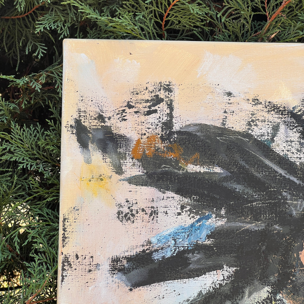 An abstract canvas painting with black, gray, beige, white, and light blue brushstrokes, photographed outdoors against green evergreen foliage.