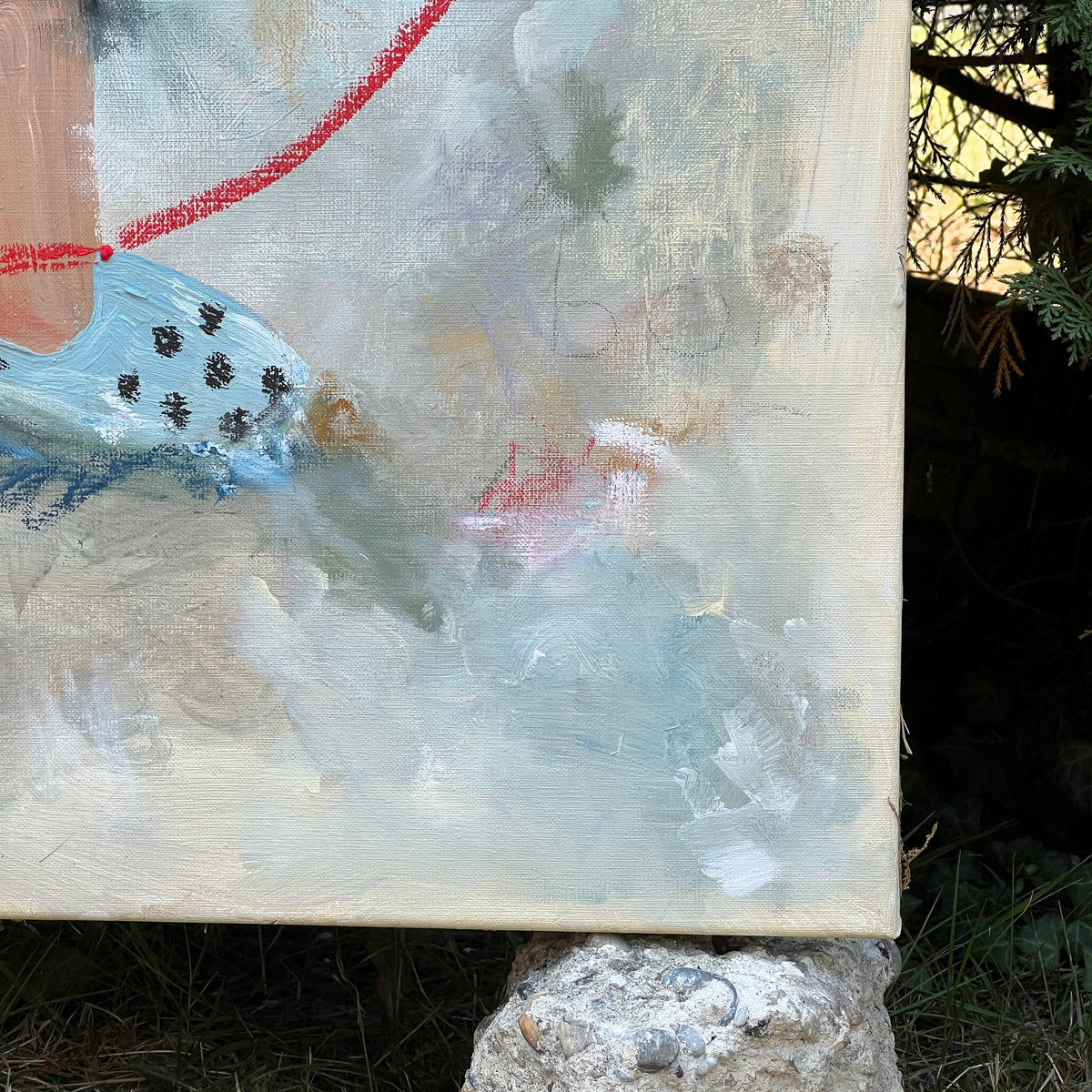 A close-up of an abstract painting on canvas, resting outdoors on a rough stone block over grass. The artwork features textured brushstrokes of cream, light blue, gray, and a bright red line.