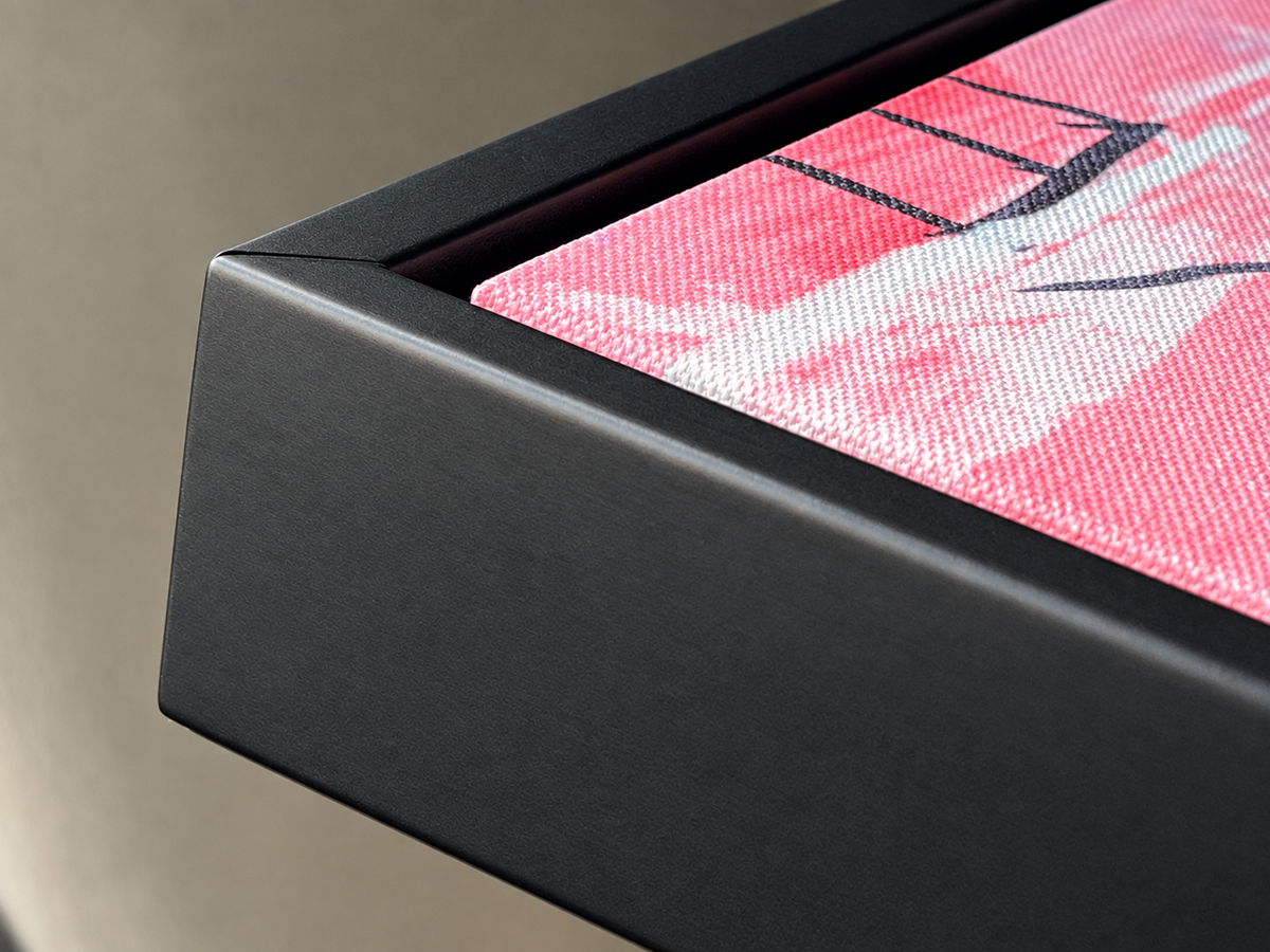 Close-up of a black rectangular frame or tray corner with a pink and white patterned textile visible inside, against a neutral beige background.